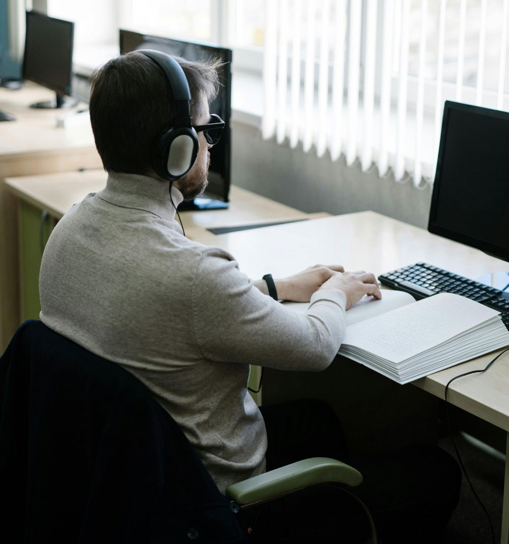 A person wearing headphones reads Braille at a computer desk in an office with several monitors